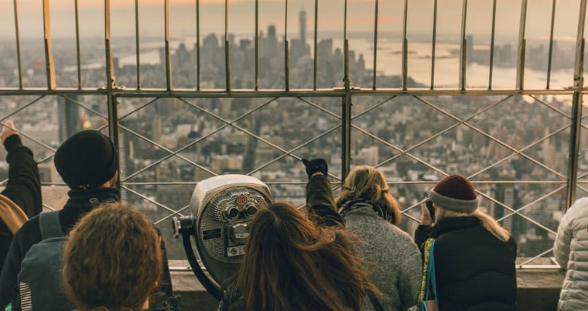 A crowd of tourists stand at the top of the Empire State Building, looking out across the Manhattan skyline