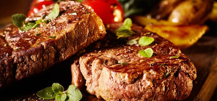 two steak cuts with leafy green garnish arranged on a wooden plank