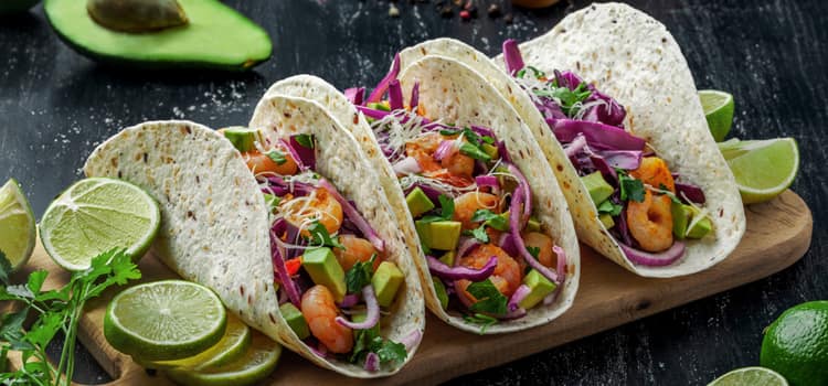 3 tacos arranged on a wooden plank filled with veggies and garnished with lime slices