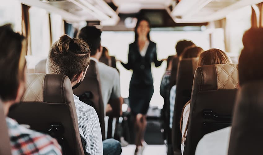 Interior of a charter bus full of travelers, a tour guide standing at the front of the cabin